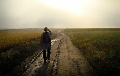 Old man with hat walking across a field on a muddy road